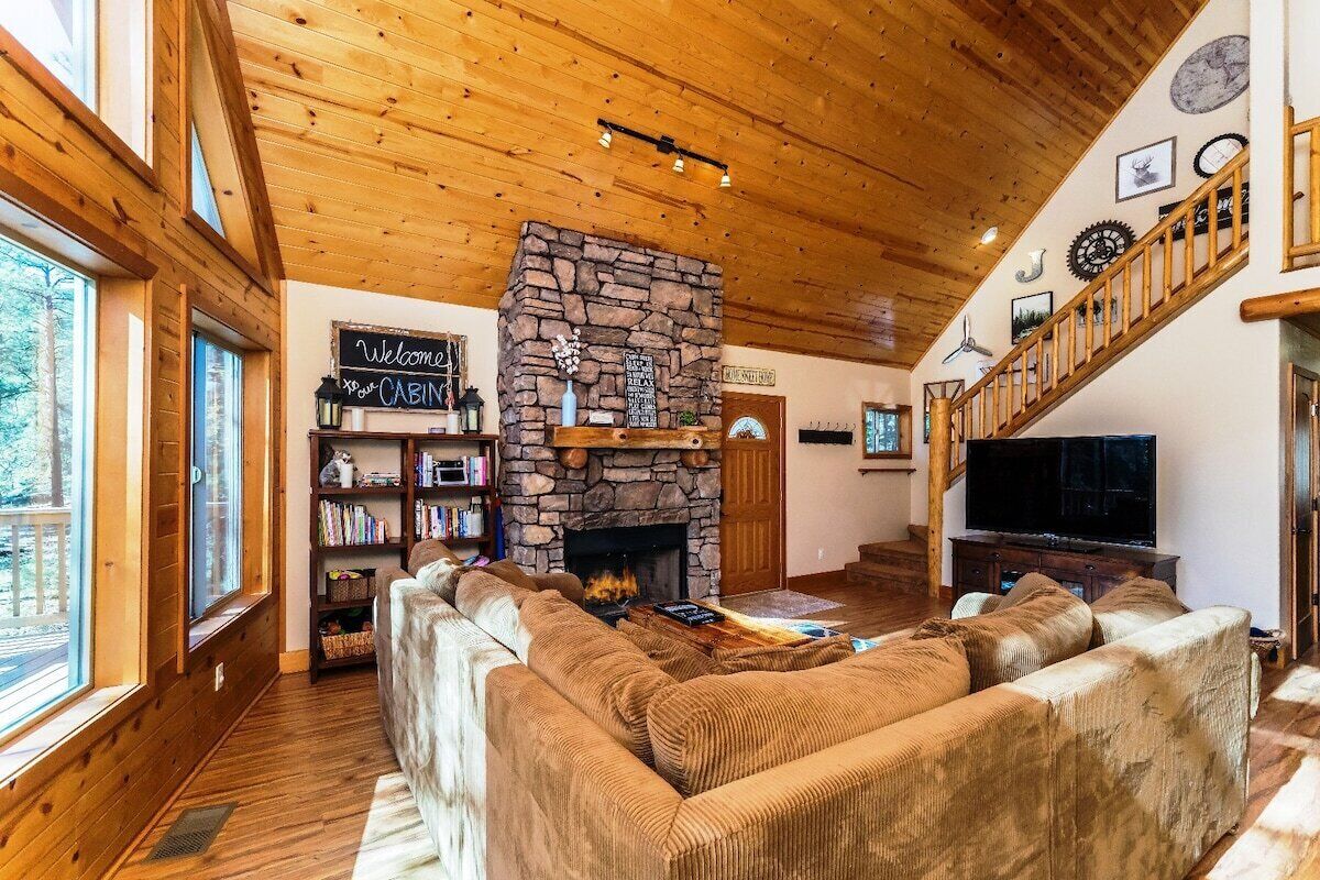 A cozy cabin interior with a stone fireplace, wooden ceiling, and a staircase.