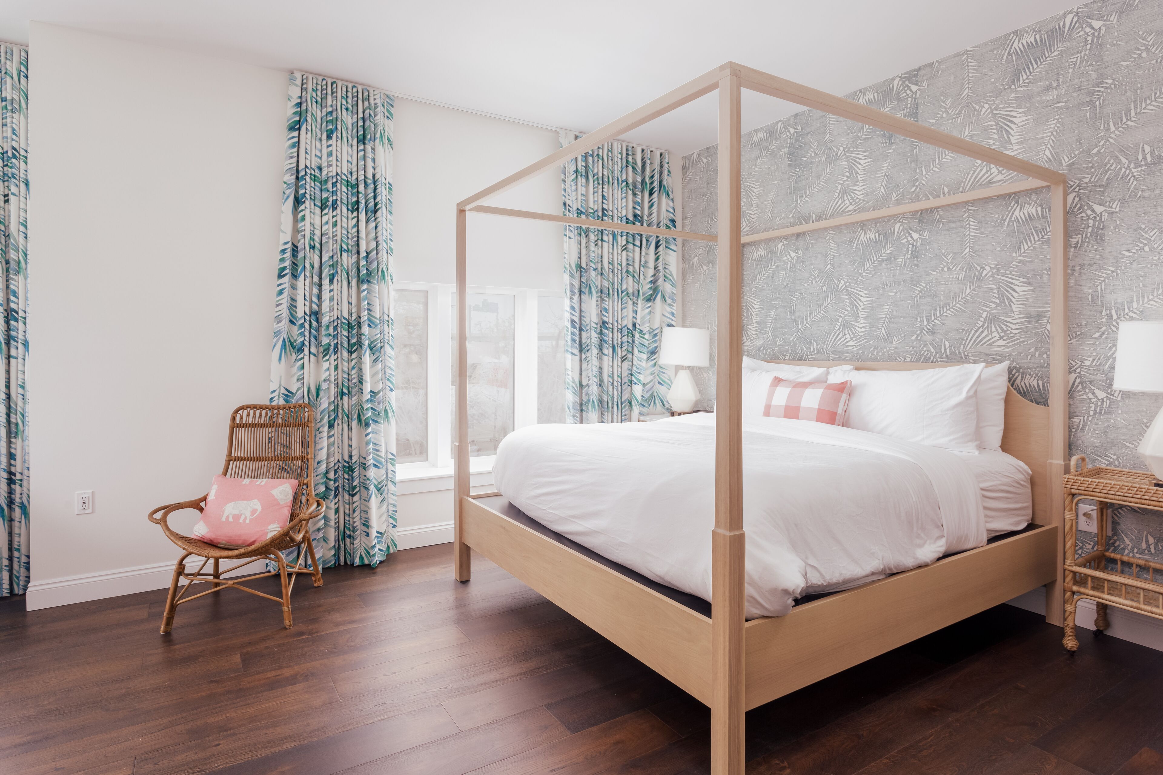 Una cama con dosel de madera, una silla de mimbre con cojín rosa y una mesita de noche con lámpara.