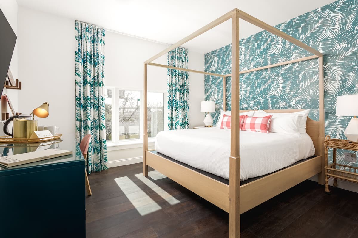 A four-poster bed with a white bedspread, a wooden headboard, and a view of the outdoors through a window with curtains.