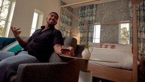 A man sitting on a couch in a modern room with a bed, a small table, and a window with curtains. - Lily Hall- A Boutique Hotel (Pensacola)