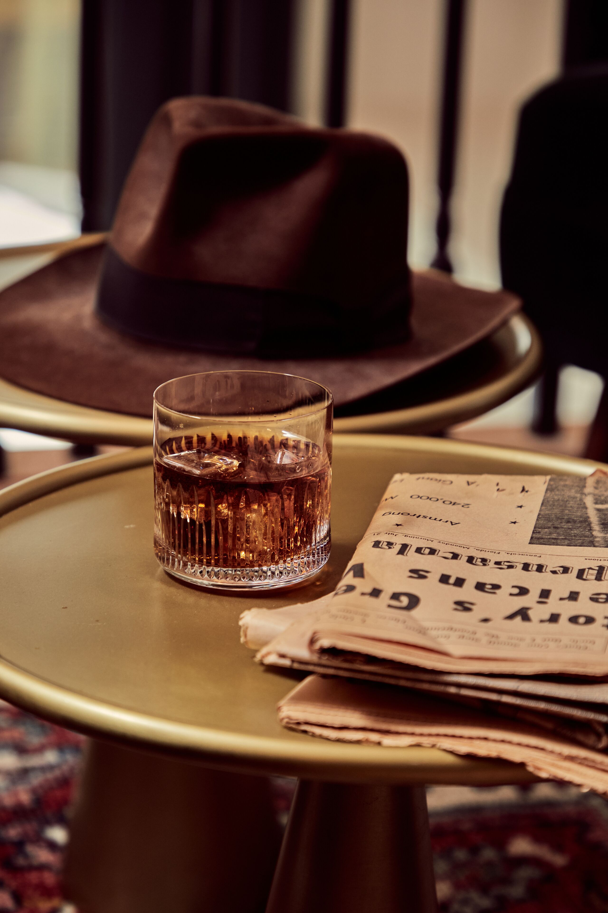 A glass of whiskey on a small round table with a newspaper and a brown hat.