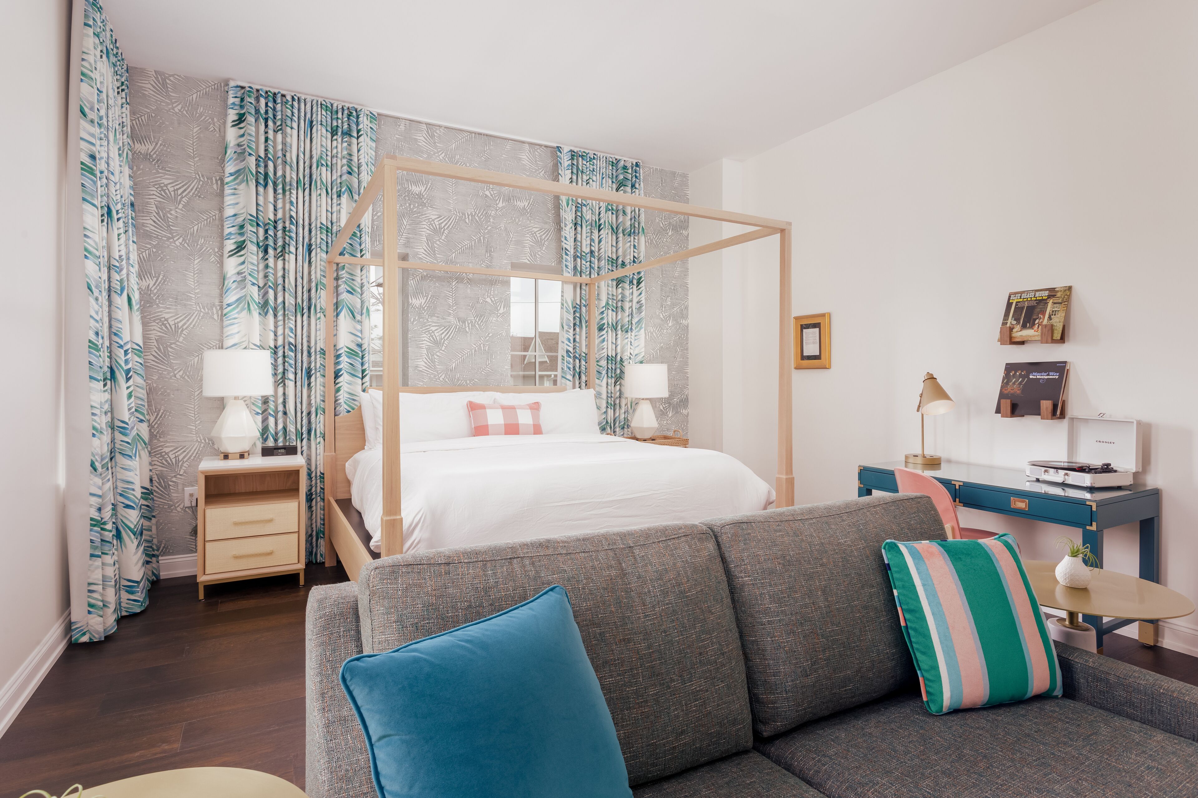 A modern hotel room with a four-poster bed, a grey sofa, and a desk with a lamp.