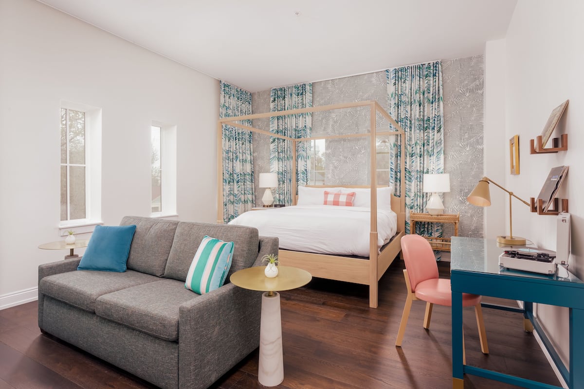 A bedroom with a four-poster bed, a gray sofa, a small round table, a desk, and a chair.
