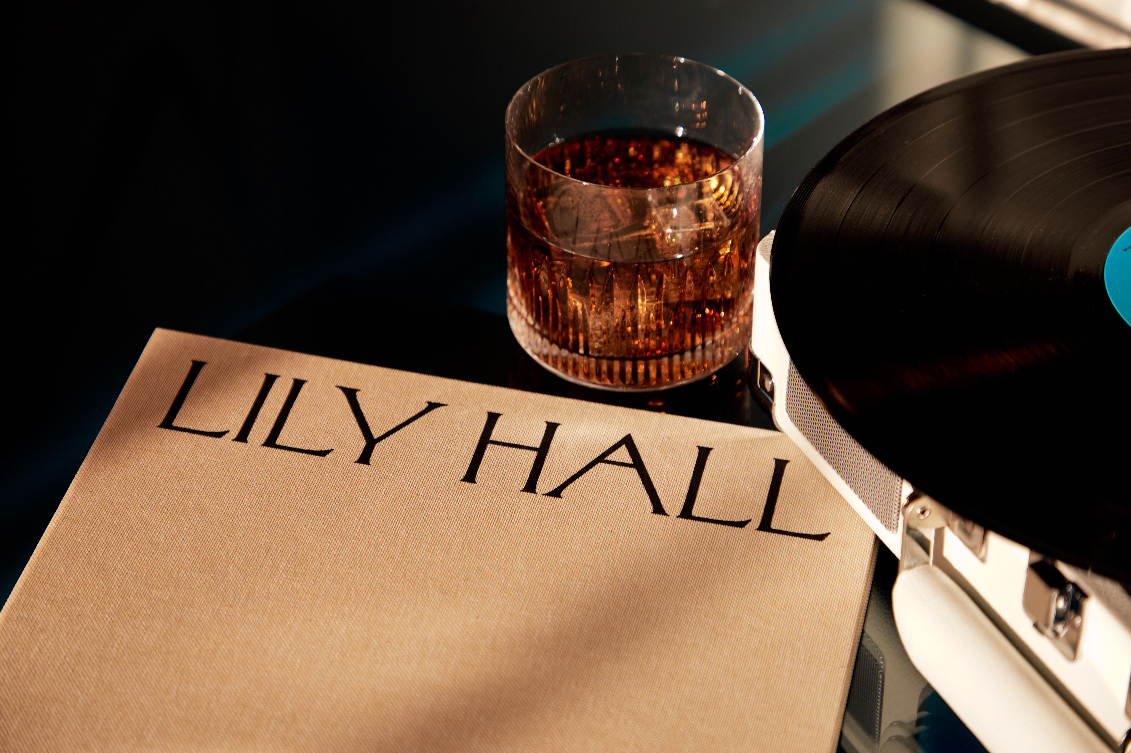 A vinyl record, a glass of amber liquid, and a beige fabric with "LILY HALL" printed on it.