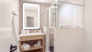 Une salle de bain avec un lavabo blanc, un miroir et une douche. On y trouve des serviettes et des articles de toilette sur le lavabo.