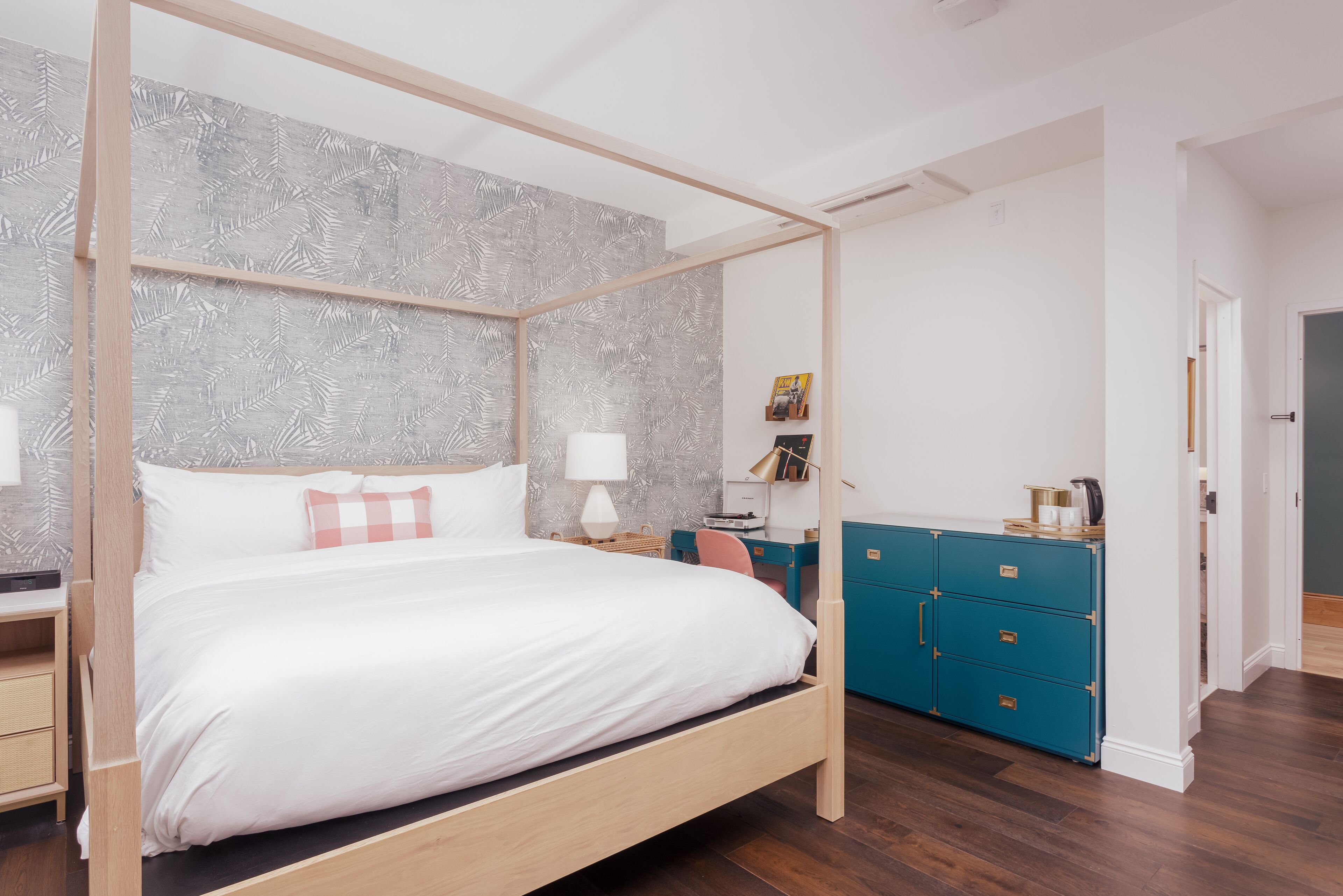 A four-poster bed with white linens, a wooden headboard, and a grey leaf-patterned wallpaper. There is a teal dresser with drawers and a small desk with a lamp.