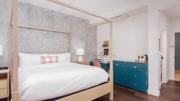 A four-poster bed with white linens, a wooden headboard, and a grey leaf-patterned wallpaper. There is a teal dresser with drawers and a small desk with a lamp.