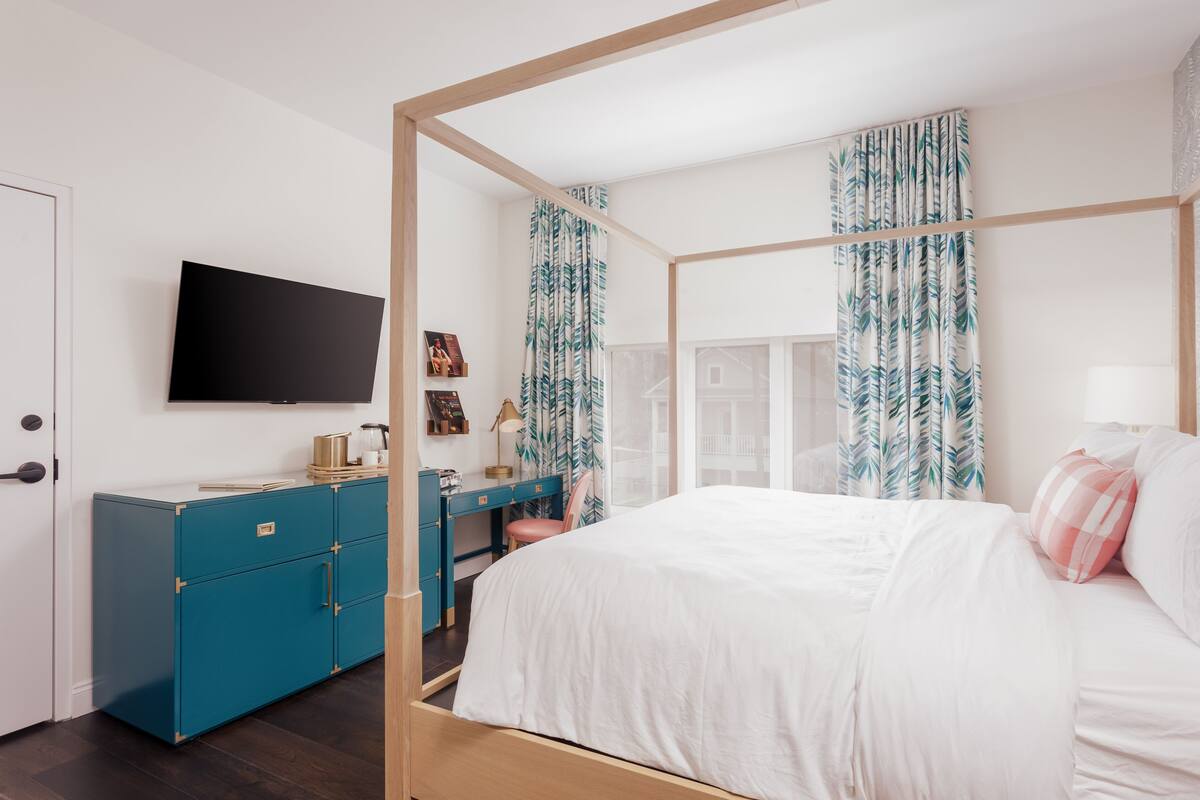 A bedroom with a four-poster bed, a desk with a chair, a television mounted on the wall, and a wardrobe.