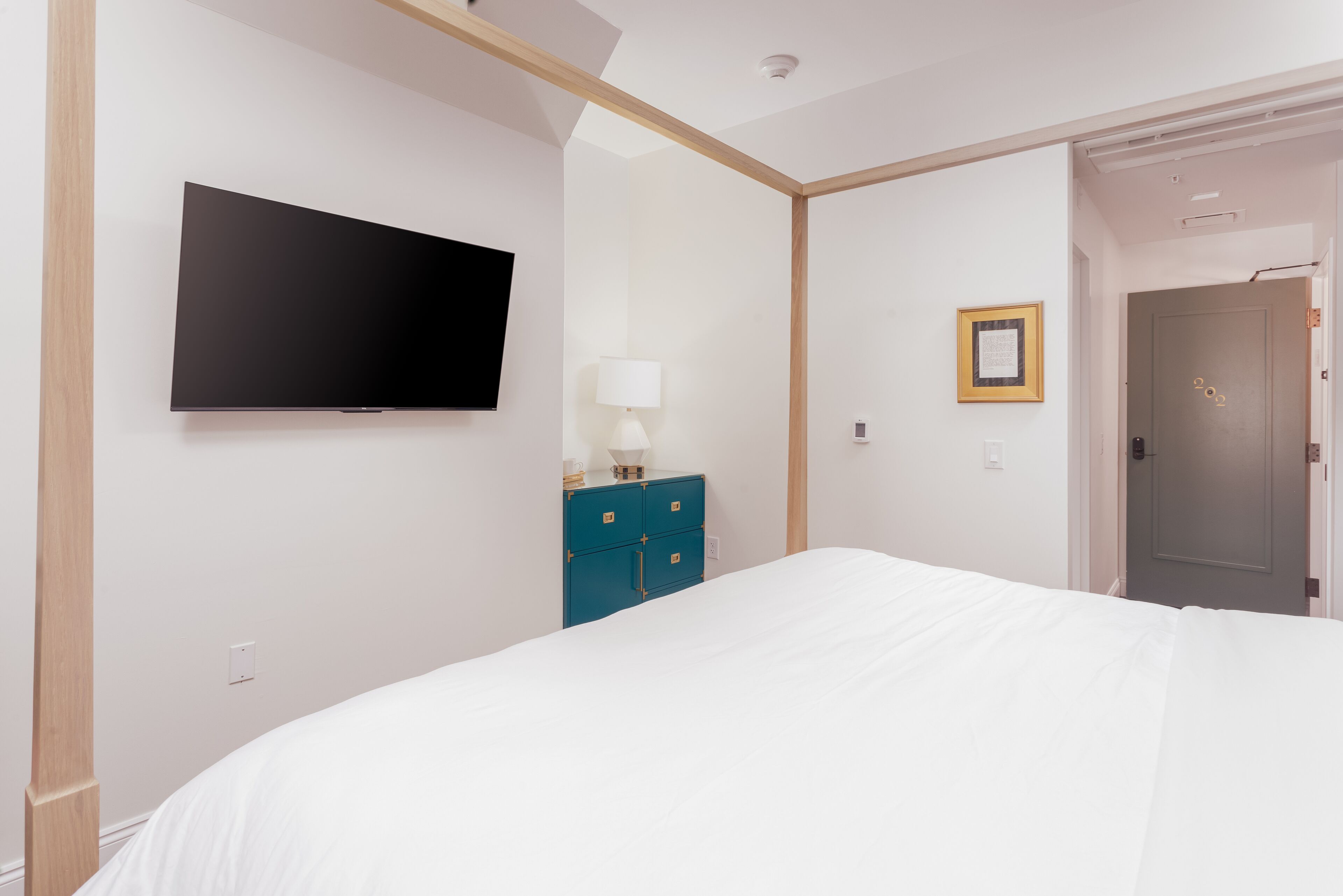 Habitación de hotel con cama, televisor de pantalla plana en la pared, cómoda, lámpara y puerta.