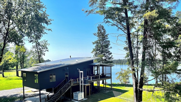 Une maison à deux étages avec un balcon donnant sur un lac, entourée d’arbres.