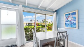 A dining area with a white table and chairs, a view of the sea through large glass doors, and a painting on the wall. - Beautiful Ground Floor 1bed/1.5 Bath Condo with Pool and Beach Access! (Siesta Key)