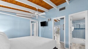 A bedroom with a bed, a ceiling fan, a TV, and a bathroom visible through an open door. - Beautiful Ground Floor 1bed/1.5 Bath Condo with Pool and Beach Access! (Siesta Key)