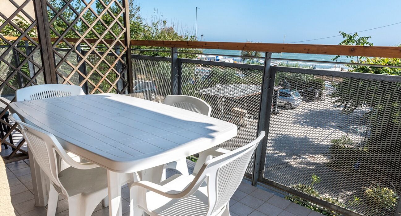 Un balcone con tavolo e sedie bianche, una recinzione a rete e vista sul mare e sugli edifici.