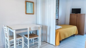 Ein Zimmer mit einem weißen Schreibtisch, zwei weißen Stühlen, einem Bett mit gelber Tagesdecke, einem Fernseher auf einer Holzkommode und einem Spiegel an der Wand.