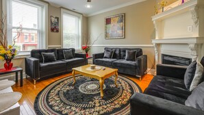 Ein Wohnzimmer mit zwei schwarzen Sofas, einem Holztisch, einem gemusterten Teppich, einem Kamin und einem Deckenventilator.