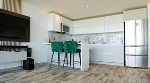 A modern kitchen with white cabinets, a stainless steel refrigerator, and a flat-screen TV mounted on the wall.