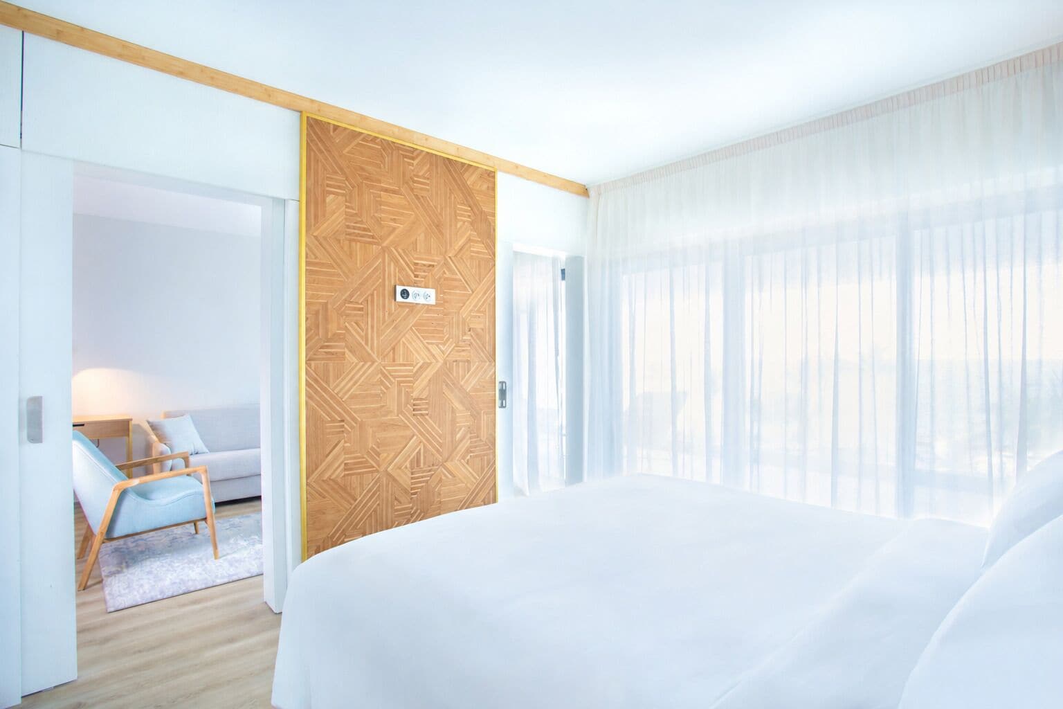 Quarto de hotel com cama, porta de madeira com padrão geométrico, janela com cortinas translúcidas e área de estar com uma cadeira e uma mesa.