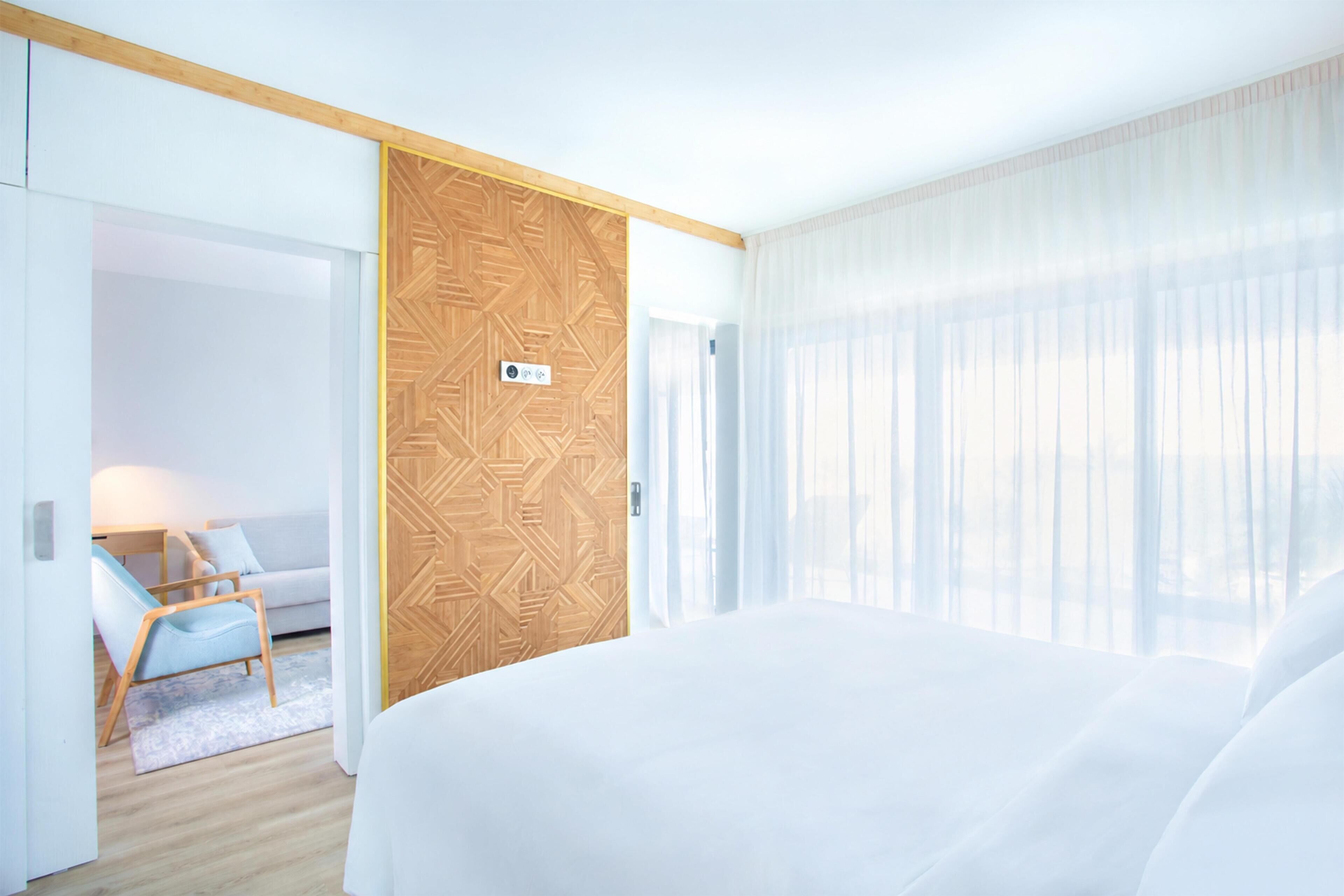 Ein Hotelzimmer mit einem großen Bett, einer Holztür mit geometrischem Muster, einer Sitzecke mit Sessel und Tisch sowie durchsichtigen Vorhängen am Fenster.