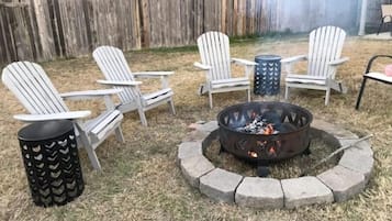 A backyard with a fire pit, four Adirondack chairs, and a metal trash can.