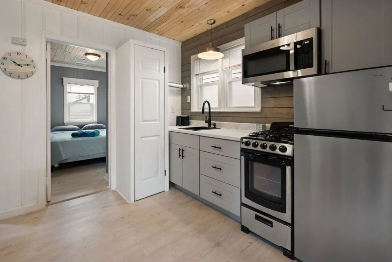 A modern kitchen with stainless steel appliances, wooden ceiling, and a view into a bedroom with a bed and window.