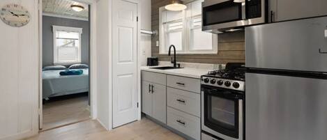 A modern kitchen with stainless steel appliances, wooden ceiling, and a view into a bedroom with a bed and window.