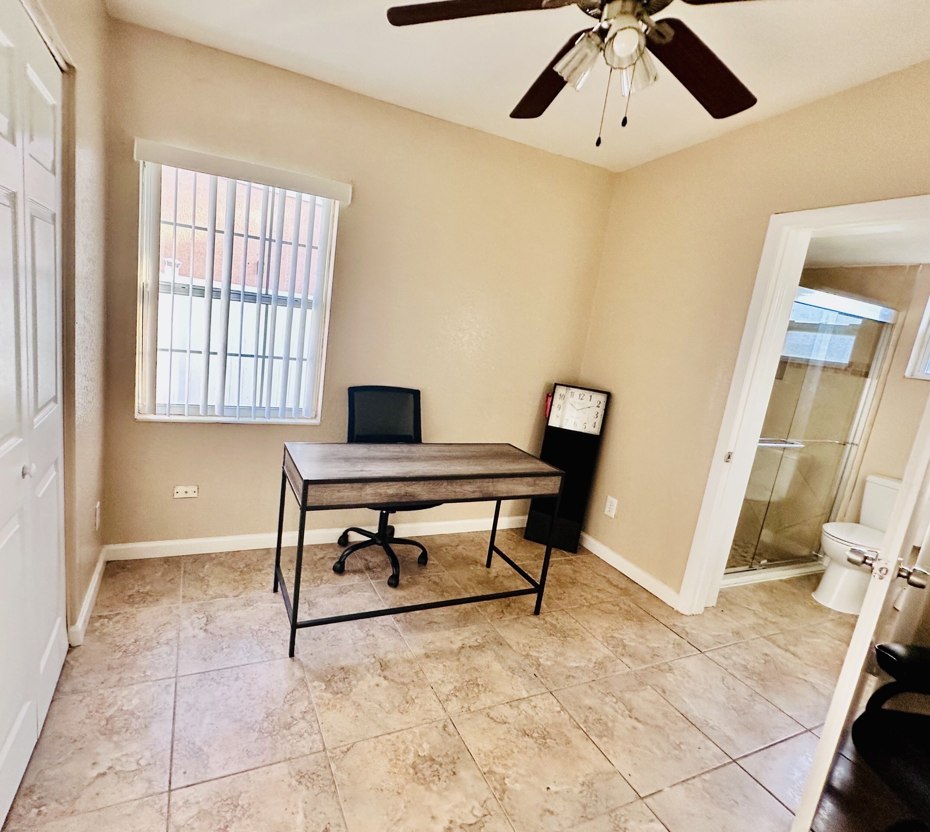 A room with a desk, chair, ceiling fan, window with blinds, and a clock on the wall.