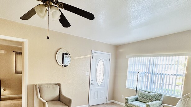 A spacious living room with a ceiling fan, a door, a window with blinds, and a patterned rug.