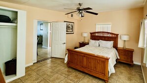 A bedroom with a wooden bed, two bedside tables with lamps, a ceiling fan, a door leading to another room, and a tiled floor.