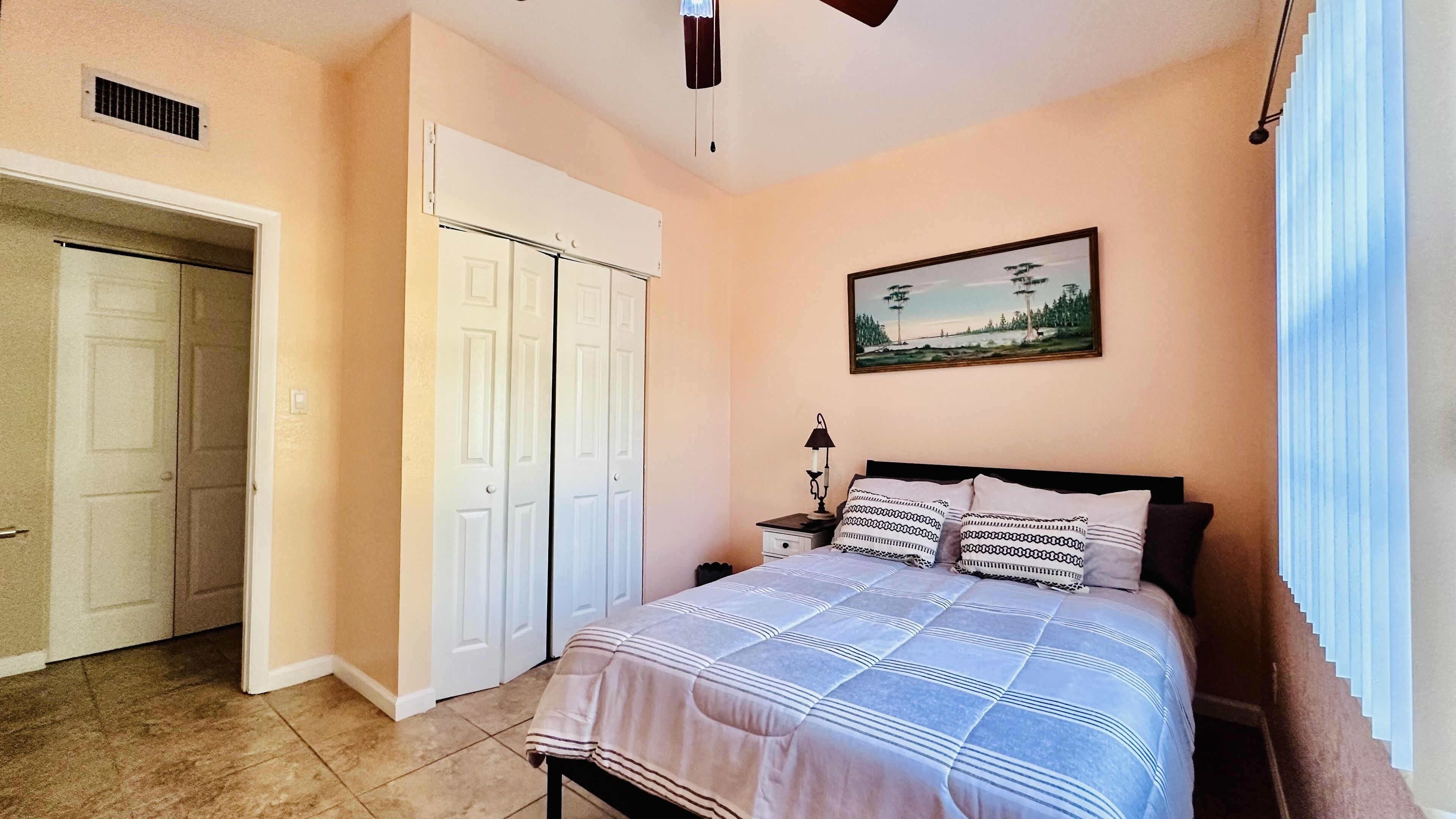 Un dormitorio con cama, mesita de noche, lámpara, un cuadro en la pared y una puerta.