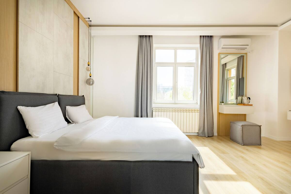 A modern bedroom with a large bed, a bedside table, a mirror, and a window with curtains.