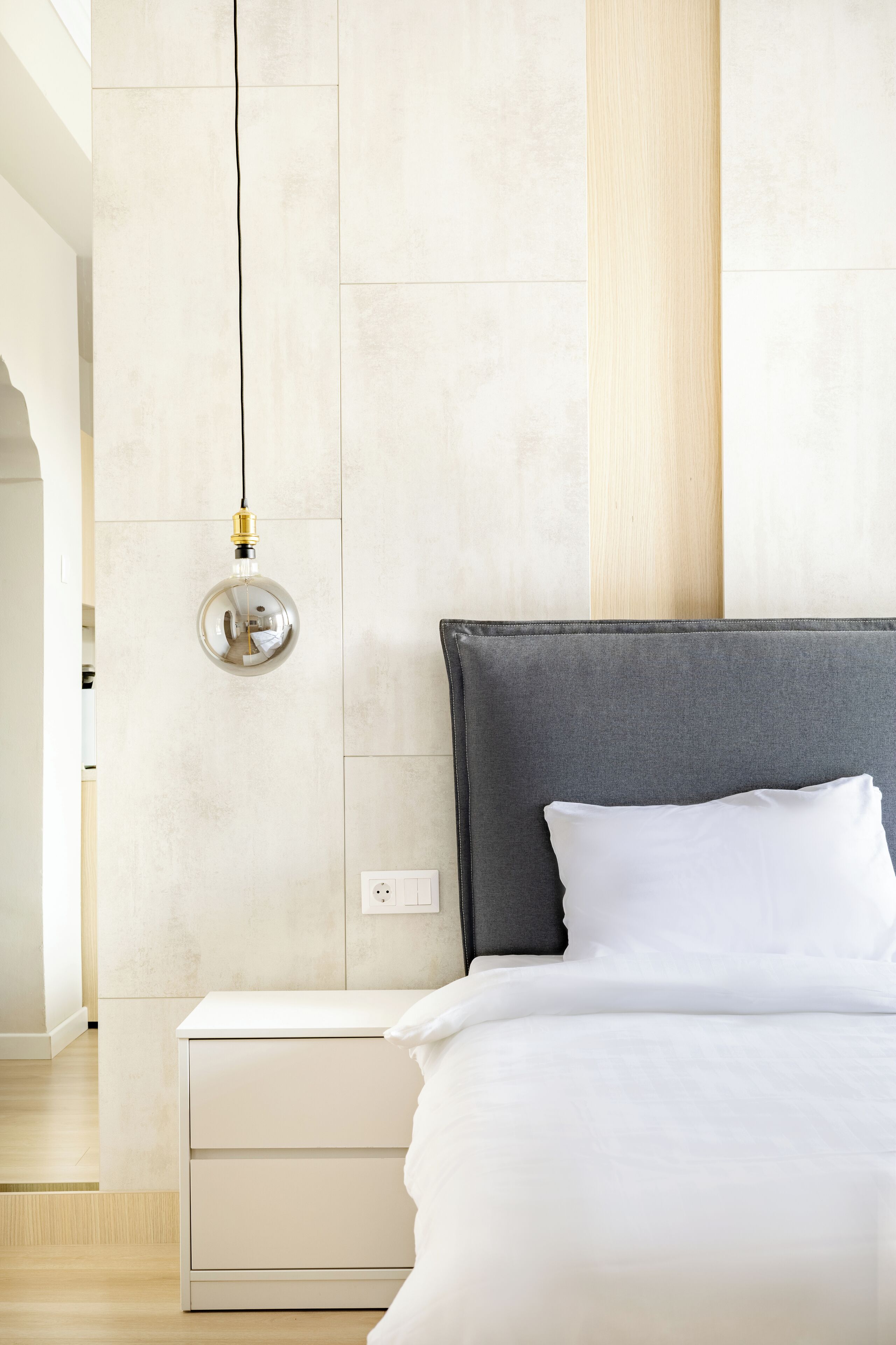 Een moderne slaapkamer met een grijs hoofdeinde, wit beddengoed en een bedlampje.