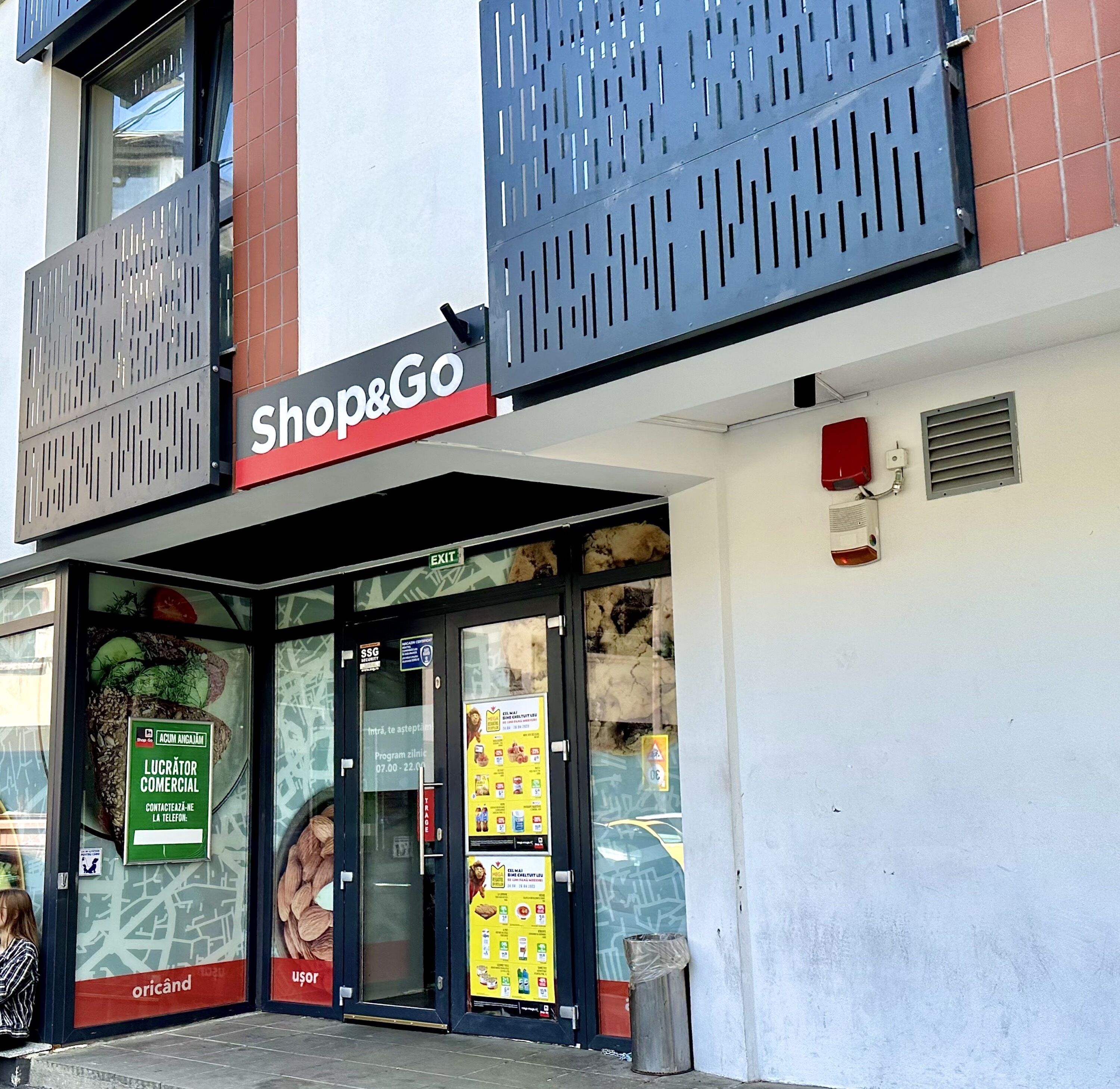 A Shop&Go store with a glass entrance, security signage, and a trash bin outside.