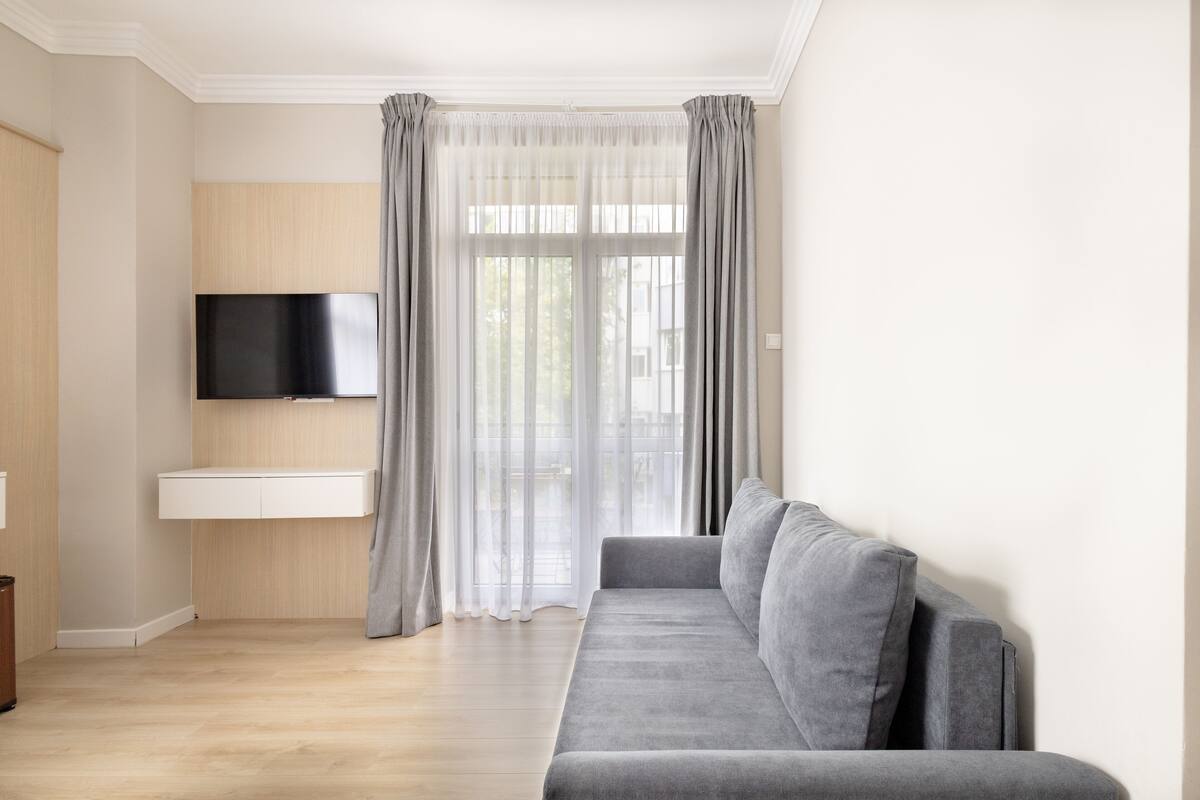 A modern living room with a grey sofa, a mounted TV, and a window with sheer curtains.
