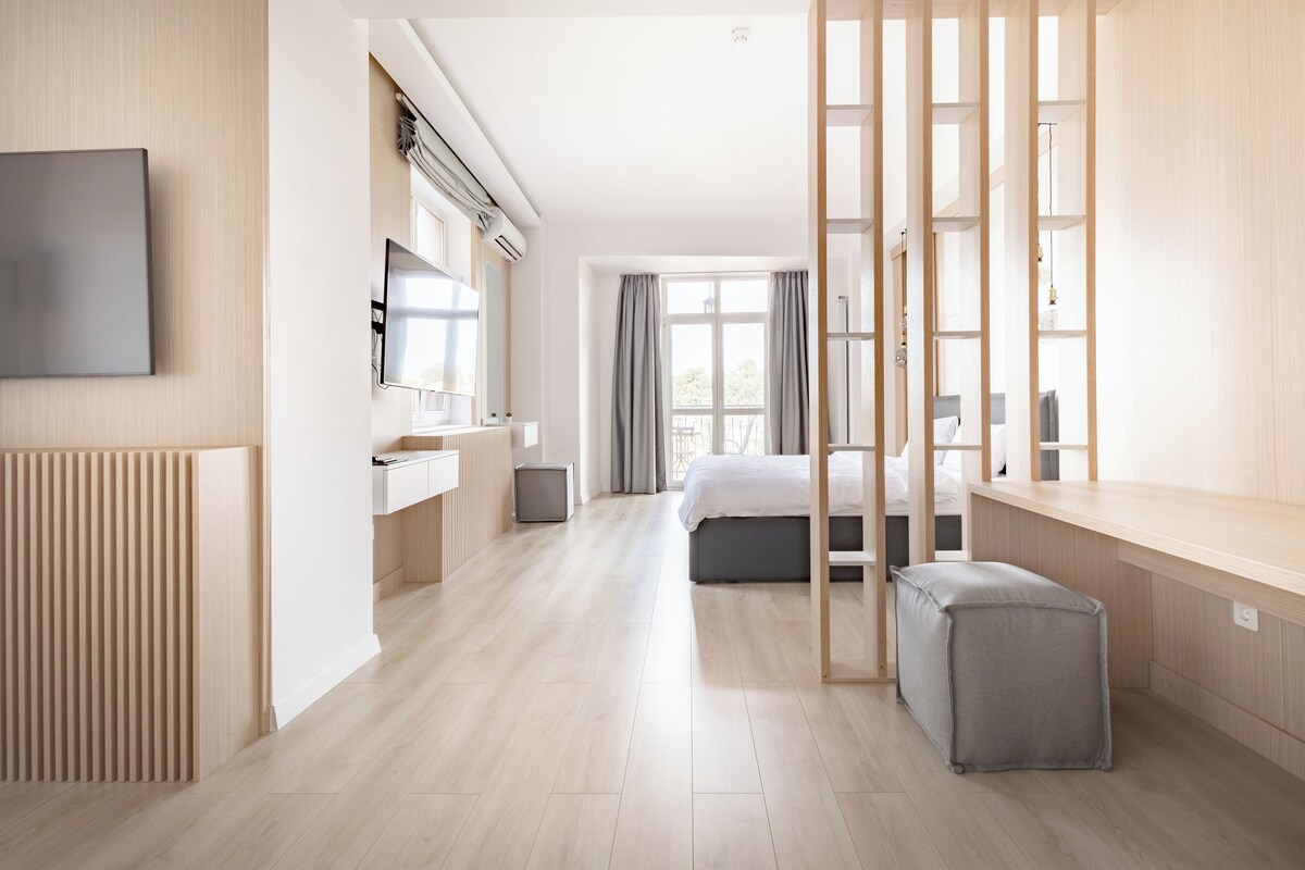 A modern hotel room with a bed, bedside tables, a TV, a desk, and a large window with curtains.