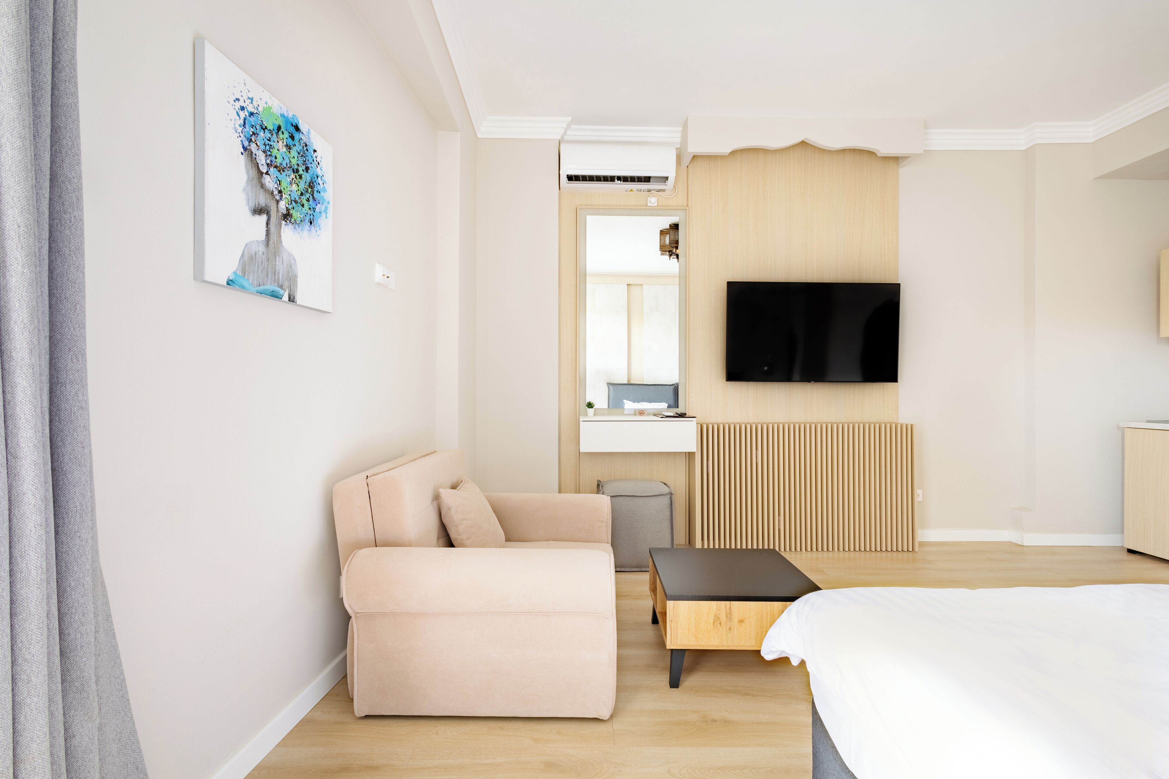 A modern hotel room with a beige sofa, a wooden coffee table, a flat-screen TV mounted on the wall, and a bed with a white duvet.
