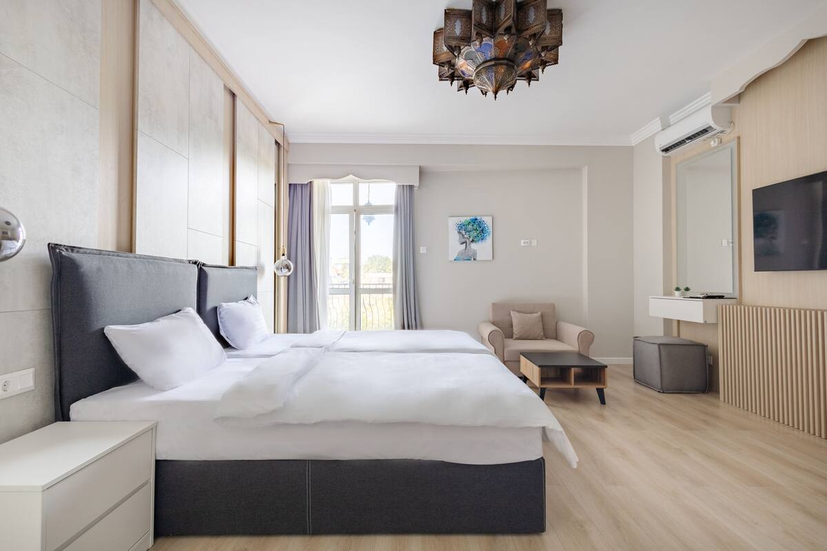 A modern hotel room with a large bed, a desk, a chair, a television, and a balcony with curtains.