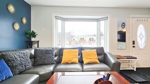 En stue med en lædersofa, to gule puder, en blå pude, et sofabord og udsigt gennem et stort vindue.