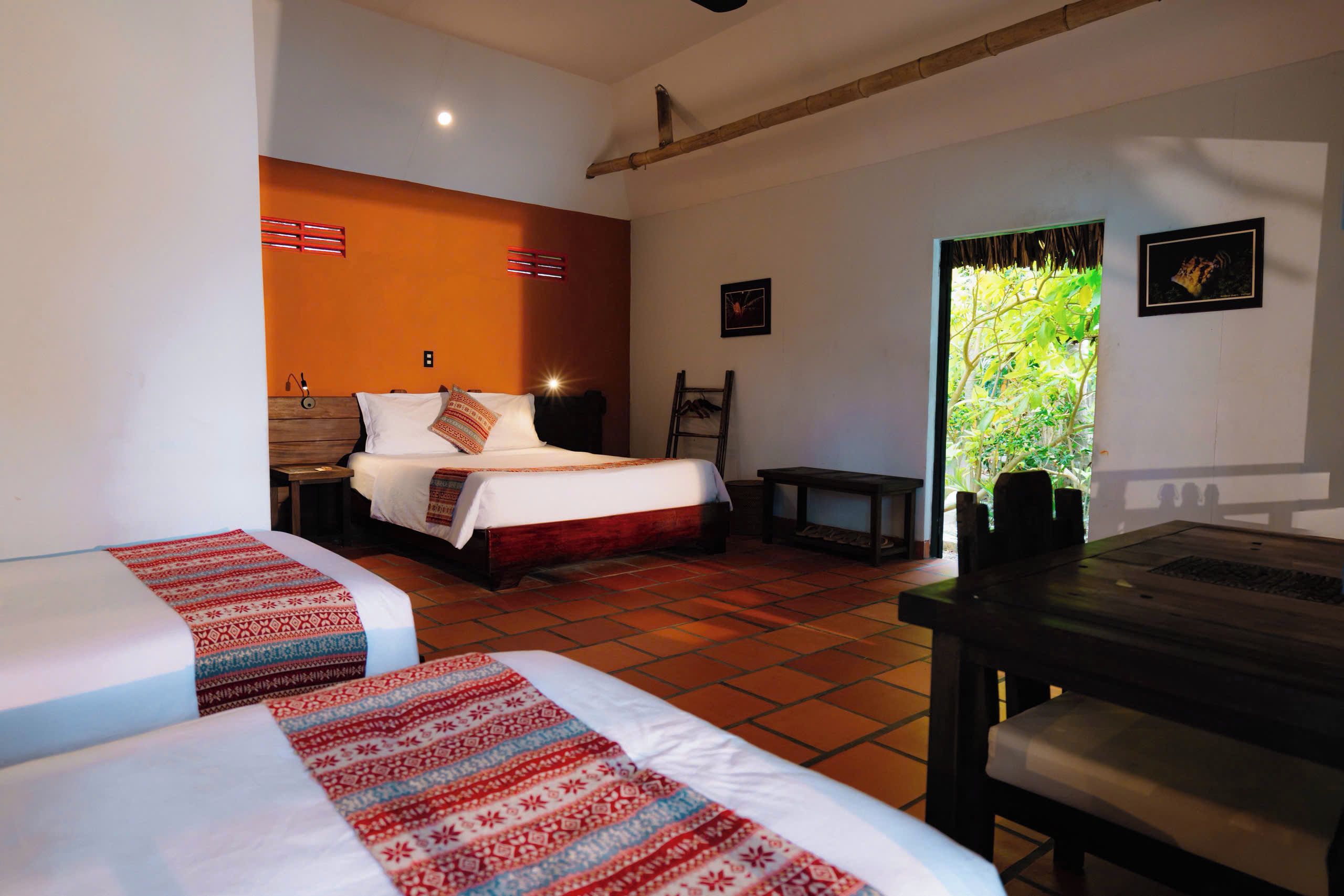 Quarto de hotel com duas camas, uma mesa de madeira, um banco, uma escada e vista para o verde pela janela.