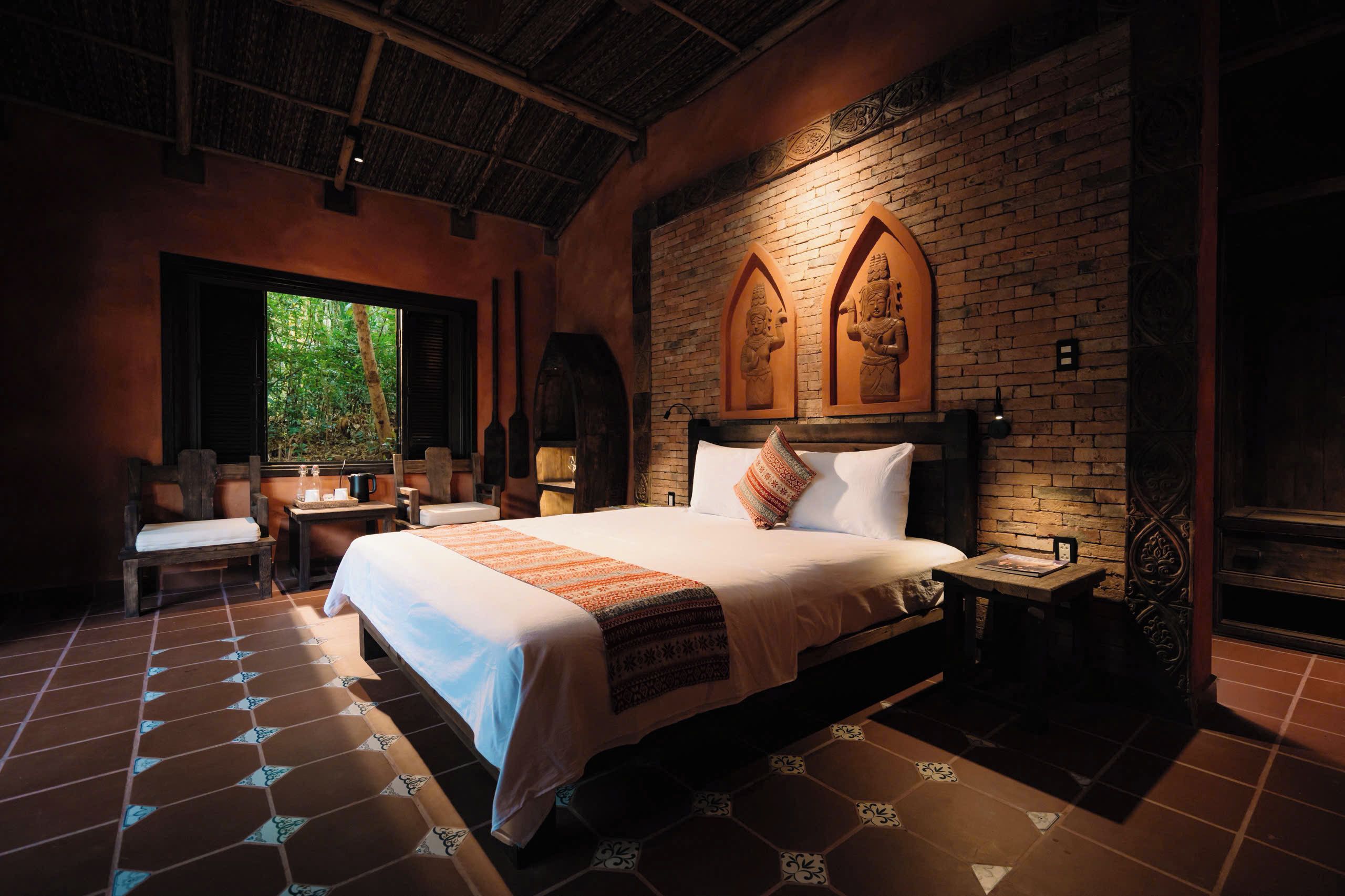 A hotel room with a large bed, a wooden chair, a small table, and a window with a view of greenery.
