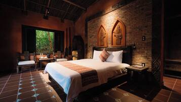 A hotel room with a large bed, a wooden chair, a small table, and a window with a view of greenery.