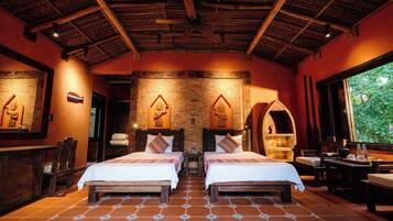 A room with two beds, a brick wall, wooden ceiling beams, and a window with a view of greenery.