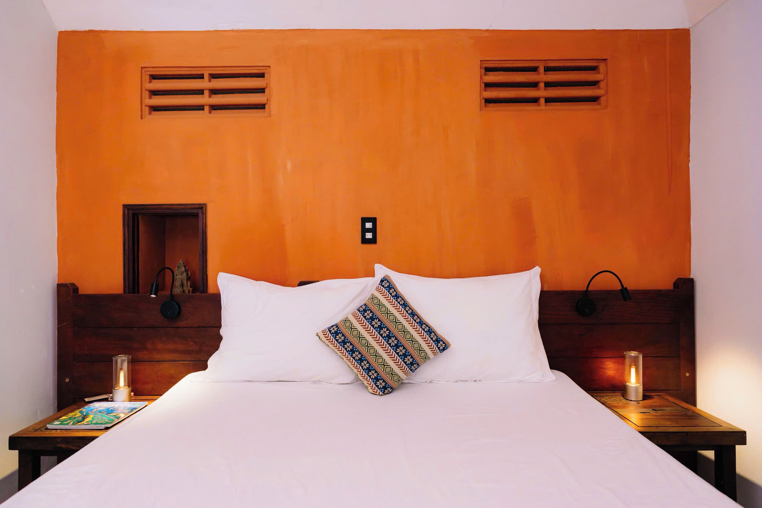Camera d'albergo con una testiera in legno, un letto bianco, un comodino con una lampada e un cuscino decorativo.