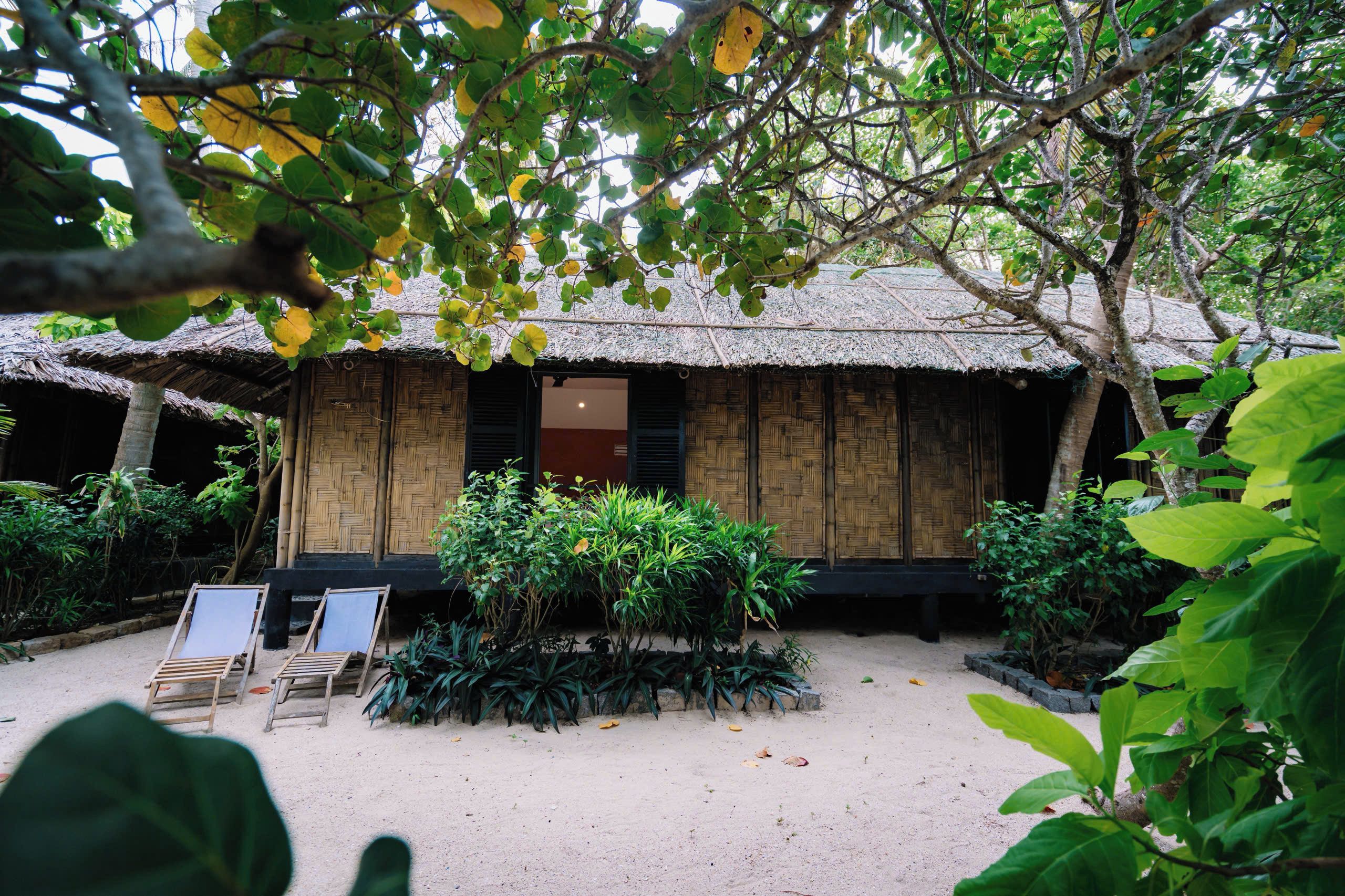 A traditional thatched-roof hut with a wooden door and windows, surrounded by lush greenery and two empty lounge chairs.