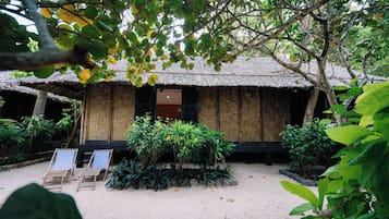 A traditional thatched-roof hut with a wooden door and windows, surrounded by lush greenery and two empty lounge chairs.