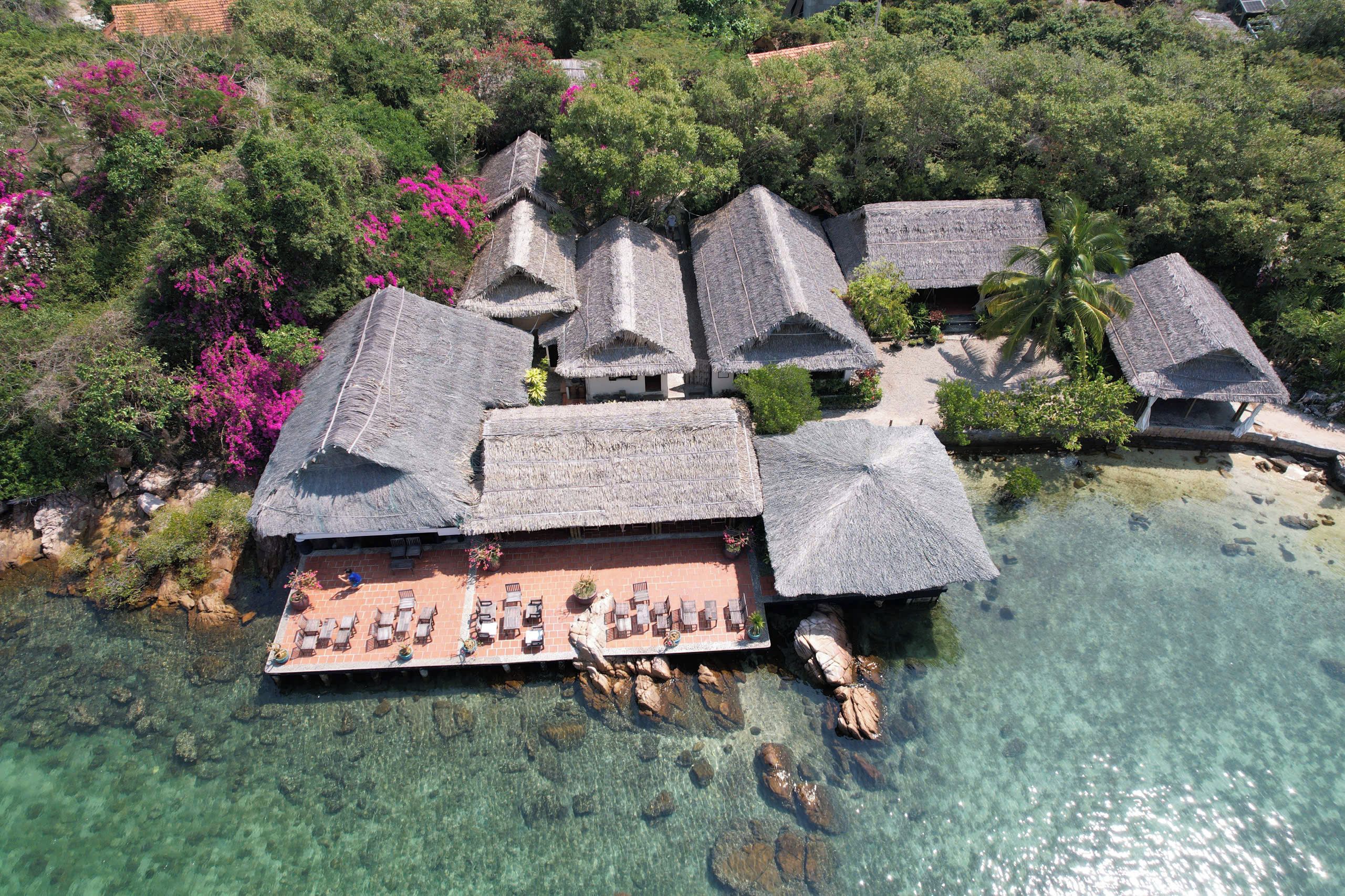 An aerial view of a resort with thatched-roof buildings, clear turquoise water, and lush greenery.