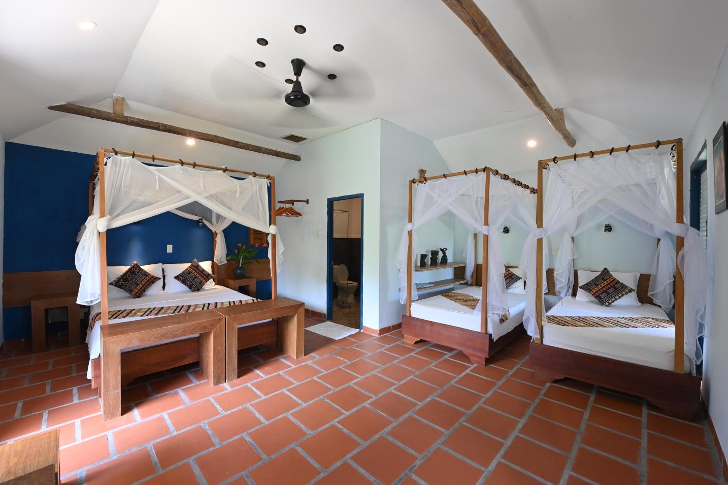 Una habitación con tres camas de madera, cada una con dosel, un banco y un ventilador de techo.