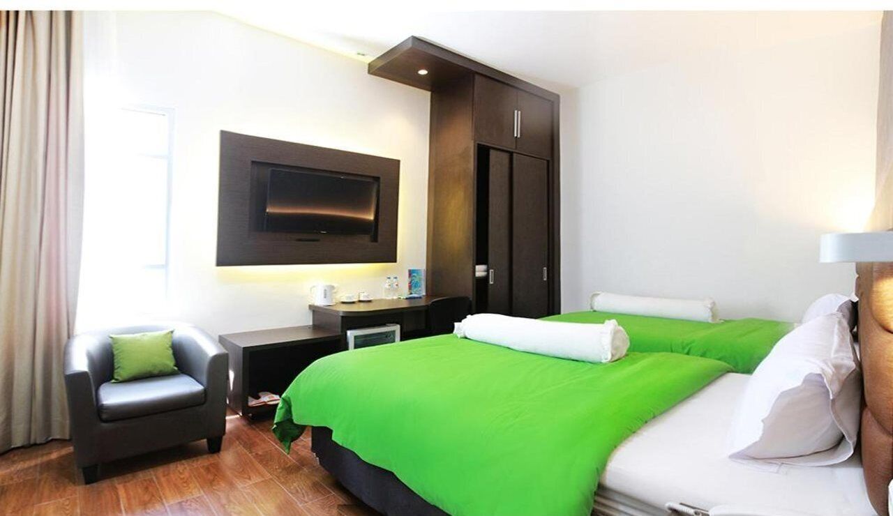 A hotel room with a bed, a desk, a chair, a TV mounted on the wall, and a window with curtains.