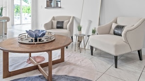 A modern living room with two beige armchairs, a wooden coffee table with a decorative tray, and a large surfboard leaning against the wall. - Wonderful Heated Pool House Delray Beach near Ocean - close to shops and the av’ (Delray Beach)