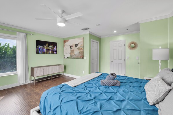 A bedroom with a large bed, a ceiling fan, a window with curtains, a wall-mounted TV, and a door.