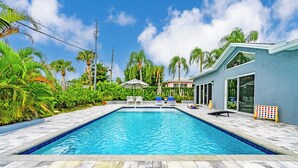 A rectangular swimming pool with a tiled deck, surrounded by palm trees and a house with large windows. - Wonderful Heated Pool House Delray Beach near Ocean - close to shops and the av’ (Delray Beach)